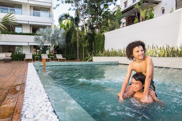 A swimming pool area with a wooden deck and a white building in the background. - Chi The Residence (Koh Samui)