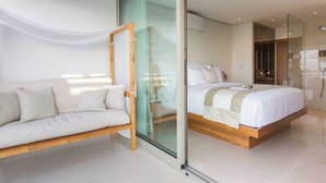 A modern bedroom with a glass door leading to a bathroom, a wooden bed frame, and a white sofa with cushions.