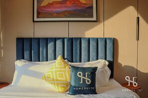 Una cama con cabecera azul, almohadas blancas y amarillas, y un cuadro enmarcado encima.
