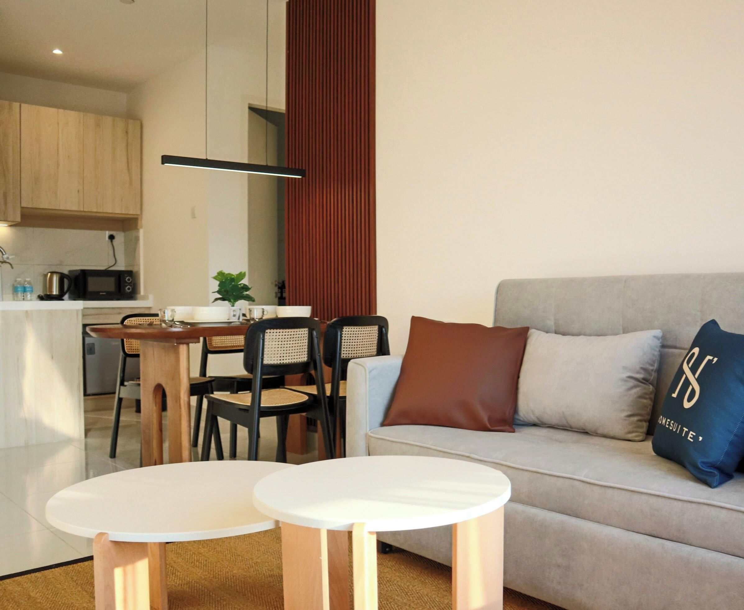 A modern living room with a sofa, two small tables, a dining area with chairs, and a kitchen area with a microwave.