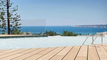 A pool with a wooden deck, a pine tree, and a view of the sea and distant buildings.