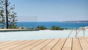 A pool with a wooden deck, a pine tree, and a view of the sea and distant buildings. - Ananea Castelo Suites Algarve (Albufeira)