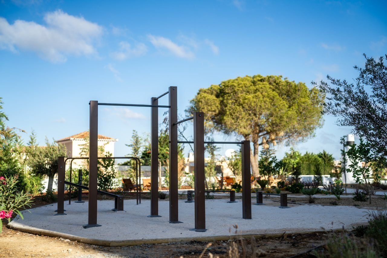An outdoor gym with exercise equipment, surrounded by a landscaped garden and a building in the background.