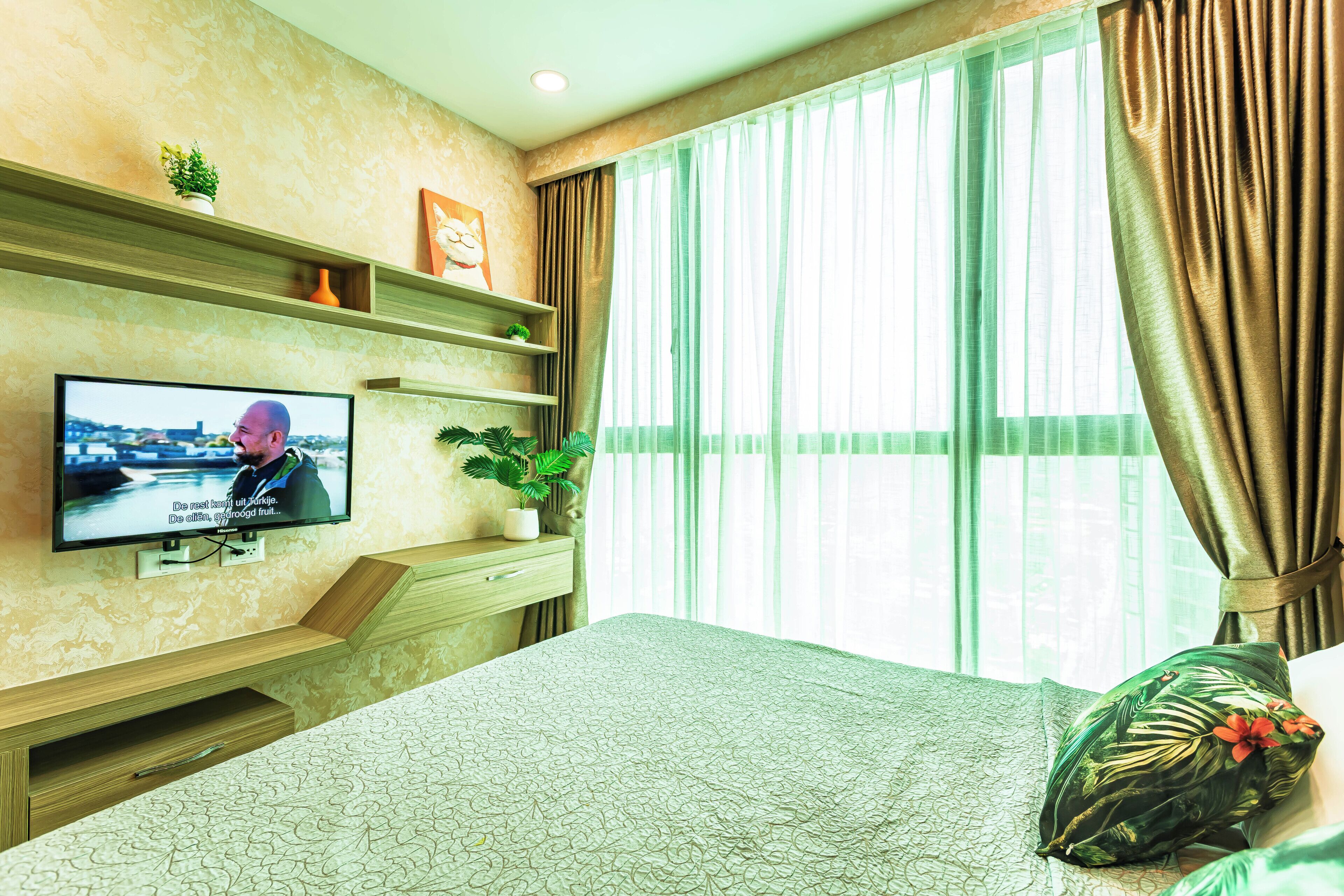 A hotel room with a television on the wall, a bed with a patterned bedspread, a potted plant, and a window with curtains.