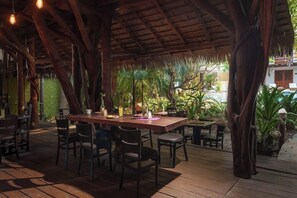 An outdoor dining area with wooden tables and chairs, surrounded by tropical plants and a thatched roof structure. - Hotel El Pueblito (Isla Holbox)