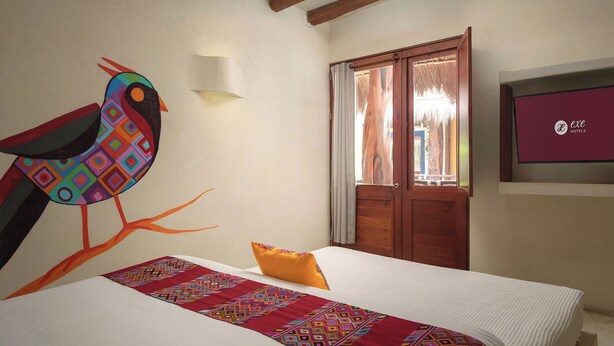 A hotel room with a bed, a colorful wall mural, a wooden door, and a TV screen displaying the hotel name. - Hotel El Pueblito (Isla Holbox)