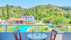 Uma varanda com duas cadeiras e uma mesa, com vista para uma piscina e uma colina com edifícios.