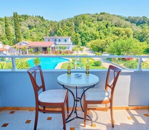 A balcony with two chairs and a table, overlooking a swimming pool and a hillside with buildings. - Sidari Green Oasis (Corfu)