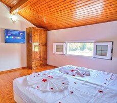 Habitación de hotel con techo de madera, cama con ropa blanca y pétalos de rosa, televisor en la pared y ventana con vistas a zonas verdes.