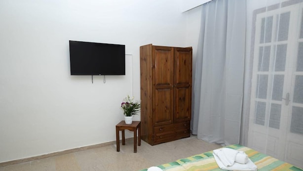 Quarto com cama, televisão, guarda-roupa, uma mesinha com planta e janela com cortinas.