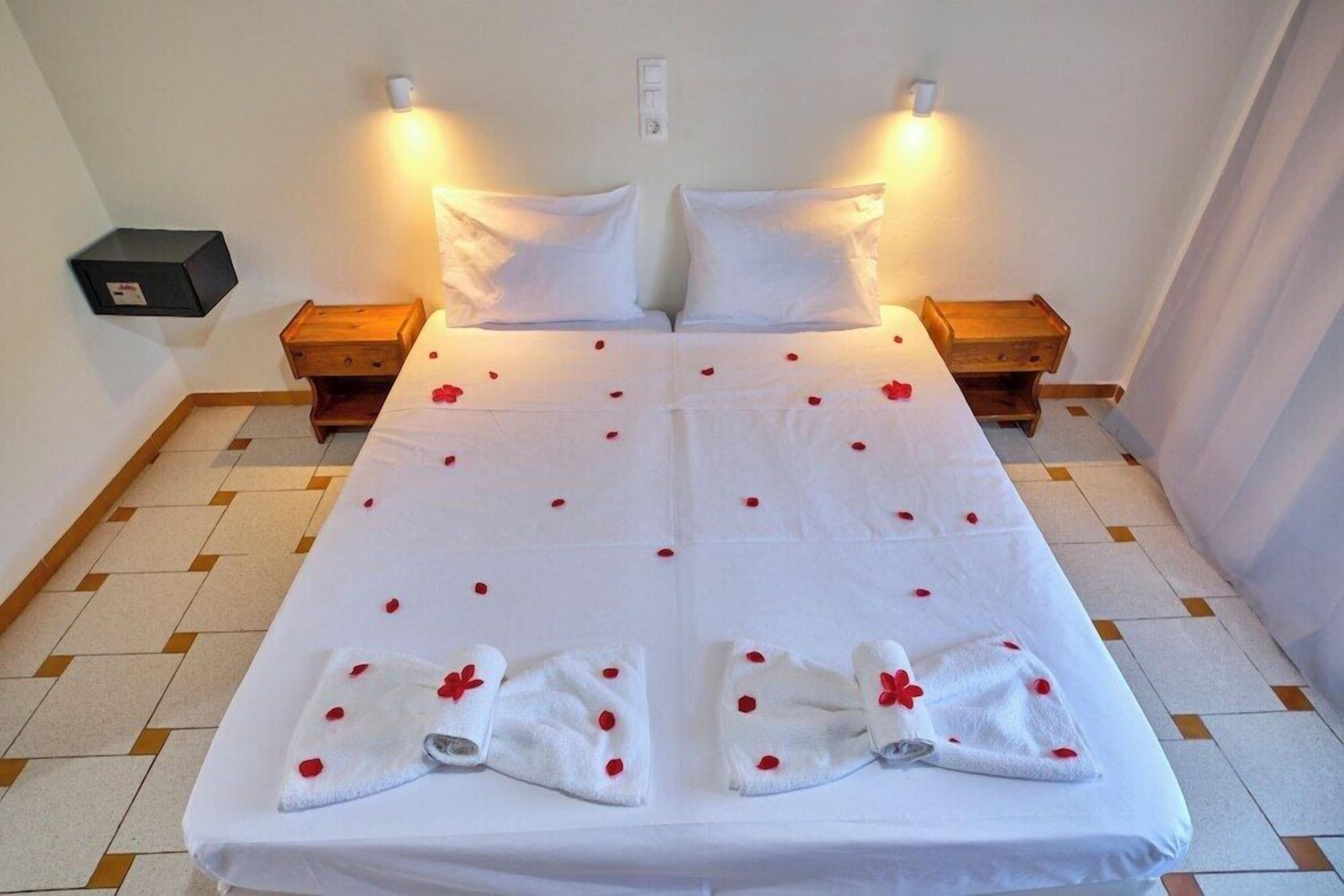 Un lit double avec du linge de lit blanc et des pétales de rose, deux tables de chevet et un luminaire fixé au mur.