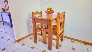 Uma mesa de jantar de madeira com cadeiras, uma tigela de frutas e um pequeno vaso de planta em um chão ladrilhado.