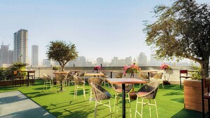 An outdoor seating area with tables and chairs, surrounded by greenery and a city skyline in the background.