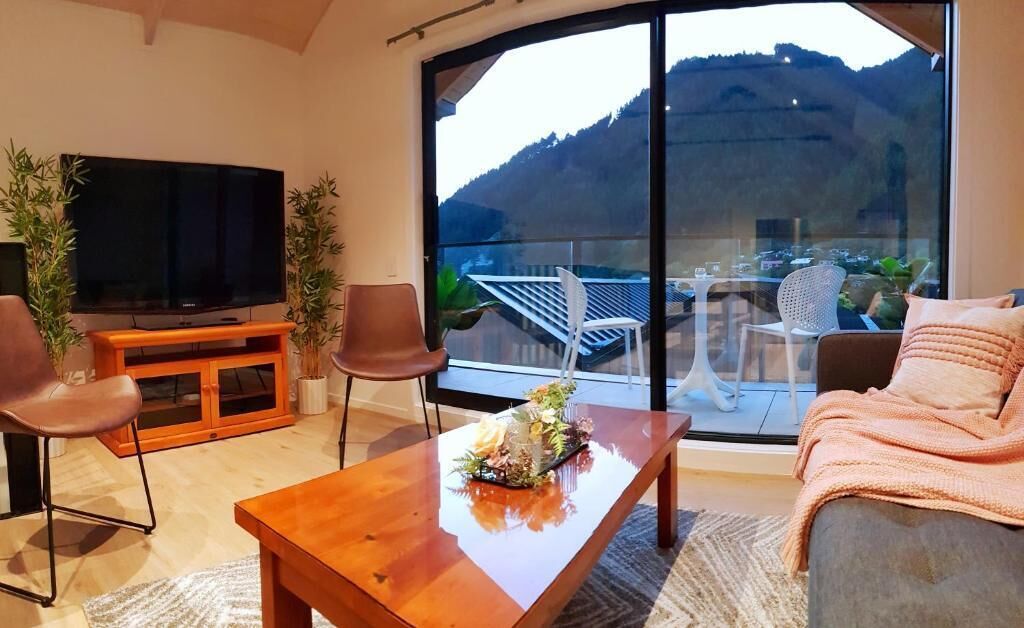 A modern living room with a flat-screen TV, a wooden coffee table, and a sofa. There is a balcony with a glass railing and outdoor seating.