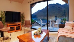 Ein modernes Wohnzimmer mit Flachbildfernseher, Holztisch und Sofa. Ein Balkon mit Glasgeländer und Sitzgelegenheit ist ebenfalls vorhanden.