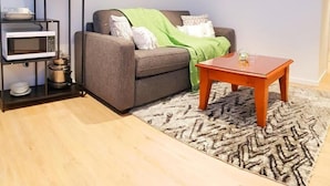 Una sala de estar con un sofá, una mesa de centro, un tapete estampado, un microondas y una estantería con utensilios de cocina.