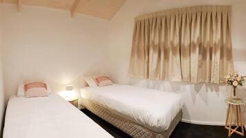 A bedroom with two beds, a bedside table with a lamp, a window with curtains, and a small table with a vase of flowers.