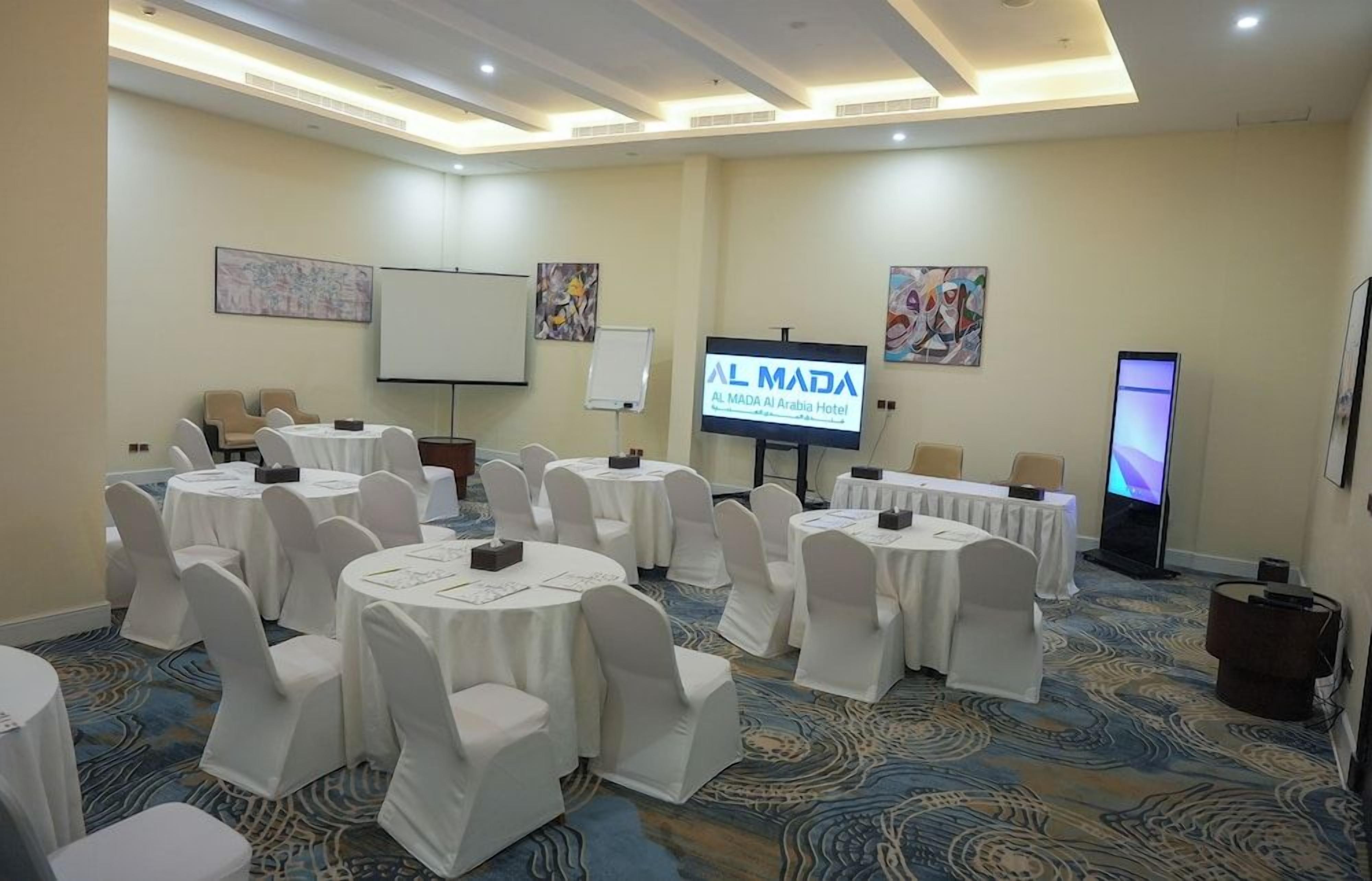 Un salón de conferencias con mesas redondas, sillas blancas, una pantalla de proyección y una televisión.
