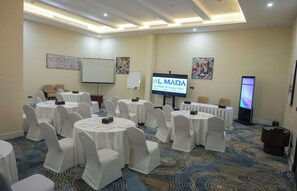 Un salón de conferencias con mesas redondas, sillas blancas, una pantalla de proyección y una televisión.