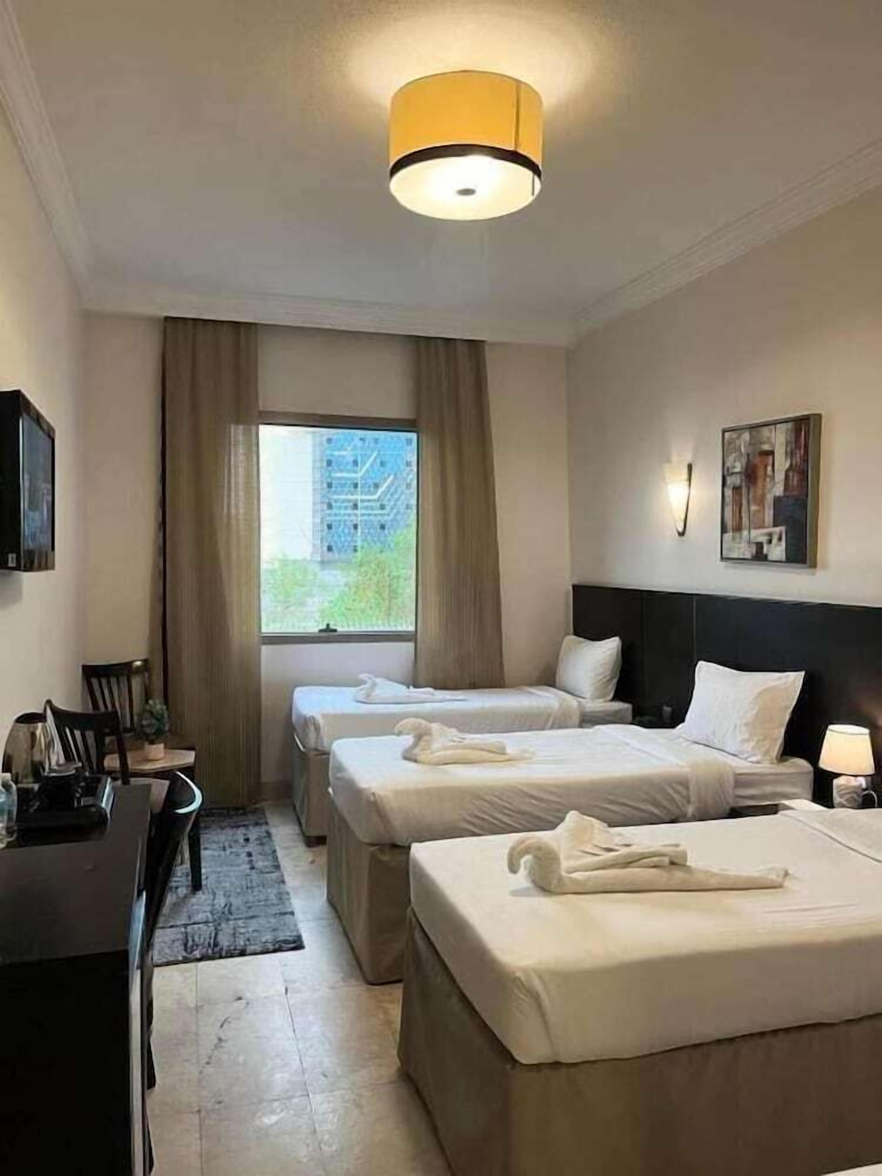 Quarto de hotel com duas camas, uma escrivaninha, uma cadeira, uma janela com cortinas, um quadro na parede e uma luminária no teto.