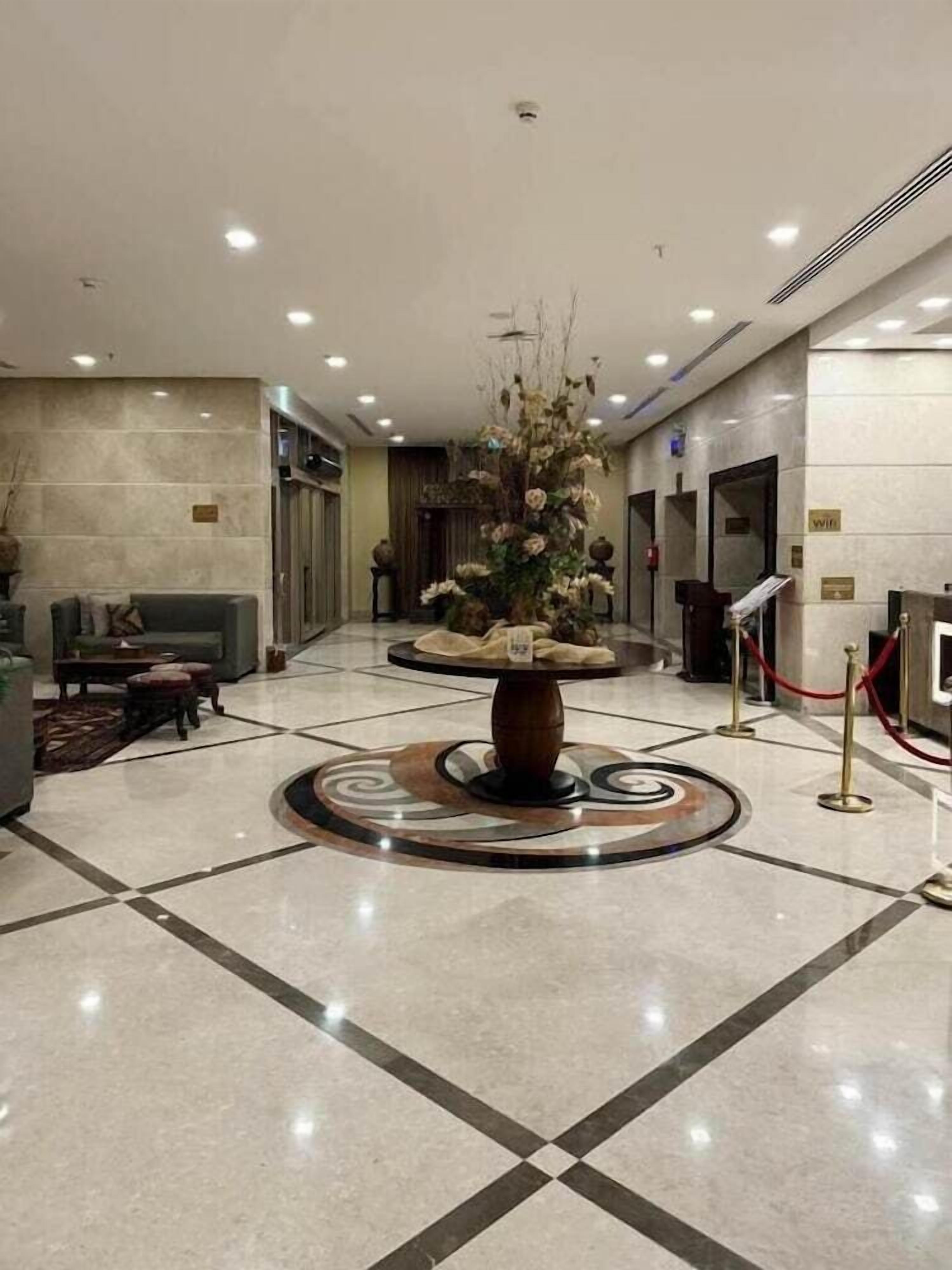 Um hall com uma mesa de recepção, área de estar com sofá e poltronas, e um vaso decorativo com flores.