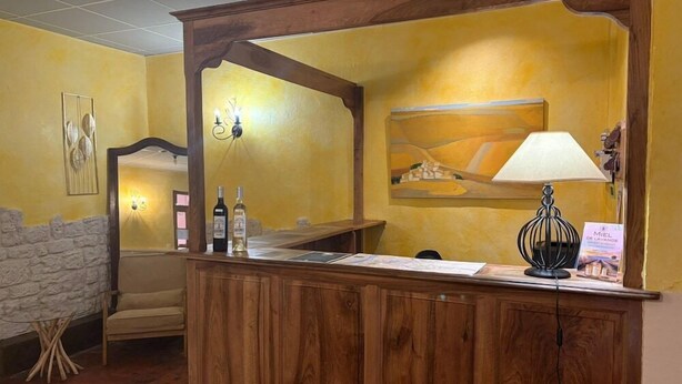 A reception desk with a wooden finish, a lamp, and a framed painting on the wall. - Adonis Digne Hotel Particulier (Digne-les-Bains)