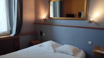 A hotel room with a bed, a mirror, a bedside table, and a window with curtains.