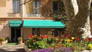 A hotel with a green awning, a flower garden in front, and a tree to the right. - Adonis Digne Hotel Particulier (Digne-les-Bains)