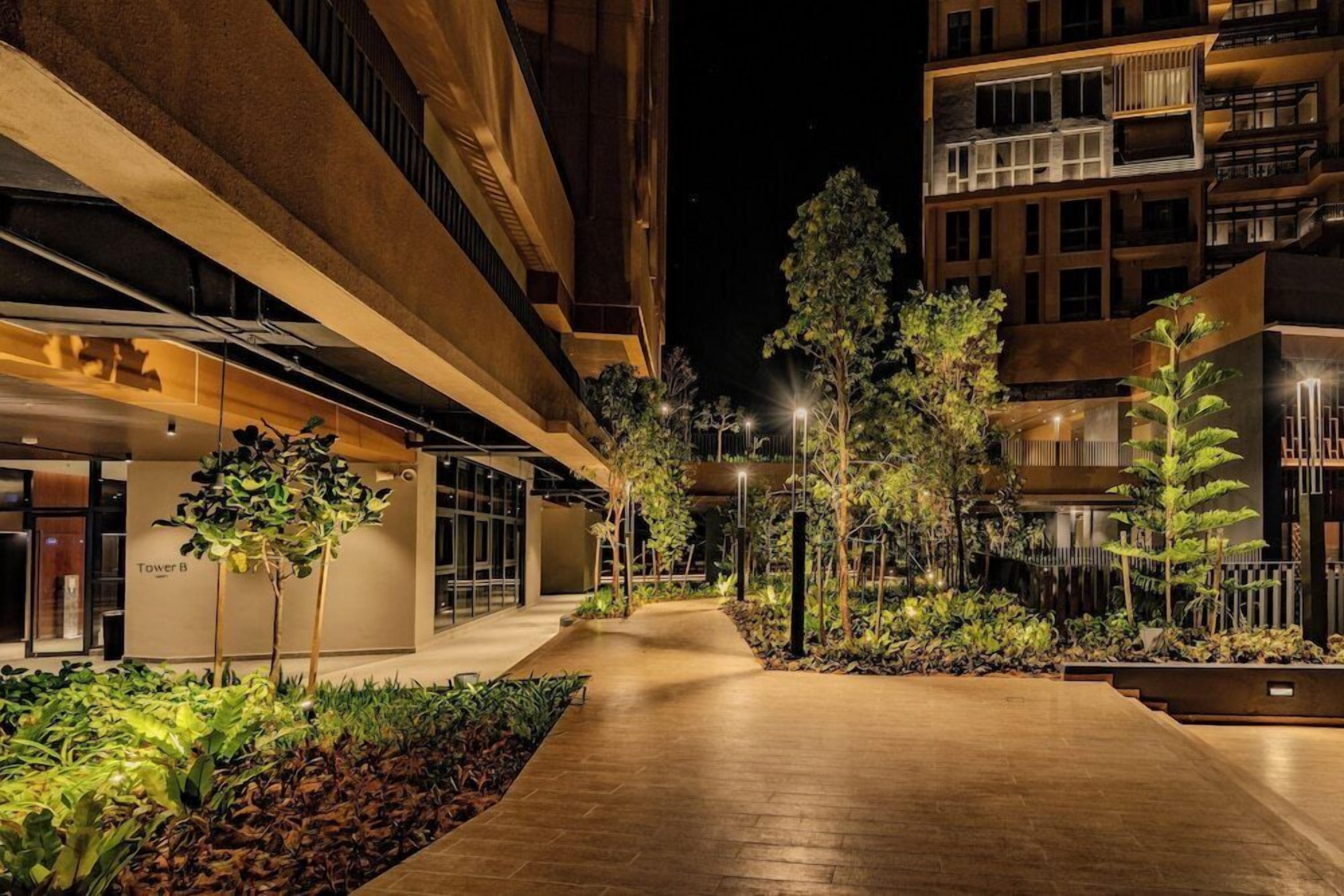 A nighttime view of a multi-story building with a central walkway, surrounded by greenery and illuminated by street lamps.