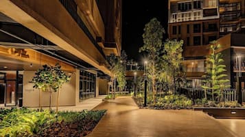 A nighttime view of a multi-story building with a central walkway, surrounded by greenery and illuminated by street lamps.