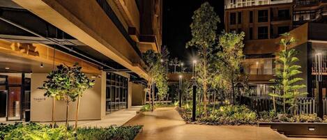 A nighttime view of a multi-story building with a central walkway, surrounded by greenery and illuminated by street lamps.
