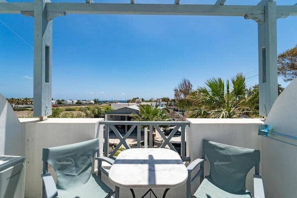 A covered outdoor seating area with a table and chairs, overlooking a coastal landscape with buildings and palm trees. - Black Coast Hotel (Santorini)
