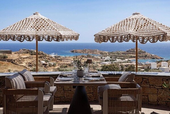A table set for two with wicker chairs and two crocheted umbrellas, overlooking a coastal landscape. - Maximus Mykonos Adults Only (Mykonos)
