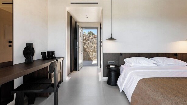 A hotel room with a bed, bedside table, lamp, and a view of a rocky landscape through an open door. - Maximus Mykonos Adults Only (Mykonos)