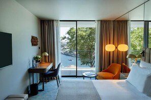 A hotel room with a bed, a desk with a chair, a small table, and a view of a body of water through large windows. - Altis Porto (Porto)