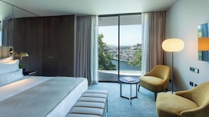 A modern hotel room with a large bed, a small round table, a sofa, and a view of a cityscape through large windows. - Altis Porto (Porto)