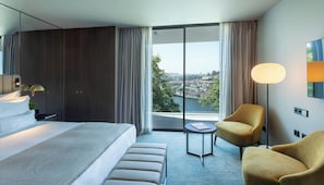A modern hotel room with a large bed, a small round table, a sofa, and a view of a cityscape through large windows. - Altis Porto (Porto)