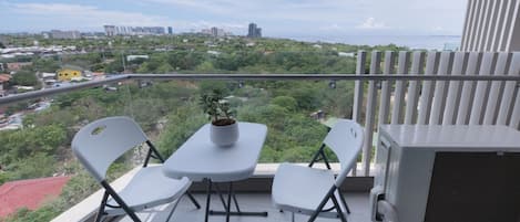 A balcony with a table and two chairs, a potted plant, and a view of a cityscape and greenery.