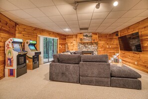 Ein gemütliches Blockhaus-Interieur mit einer Eckcouch, zwei Spielkonsolen, einem Kamin aus Naturstein und einem großen Fernsehbildschirm.