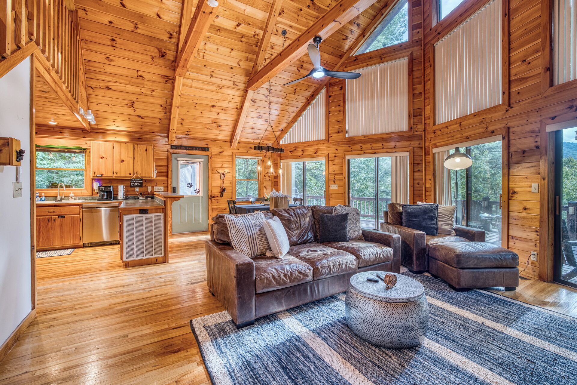 A cabin interior with a wooden ceiling, a kitchen area, a seating area with a sofa and armchairs, and large windows.