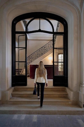A woman walking through a grand entrance with a staircase leading to an upper level.
