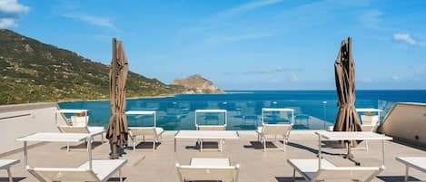 A rooftop pool area with lounge chairs and umbrellas, overlooking a clear blue sea and a hilly landscape.