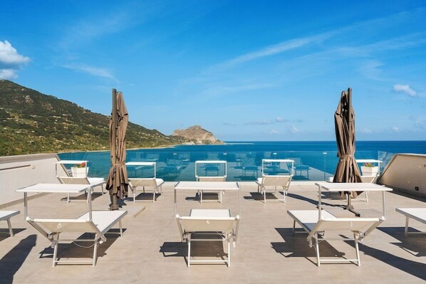 A rooftop pool area with lounge chairs and umbrellas, overlooking a clear blue sea and a hilly landscape.