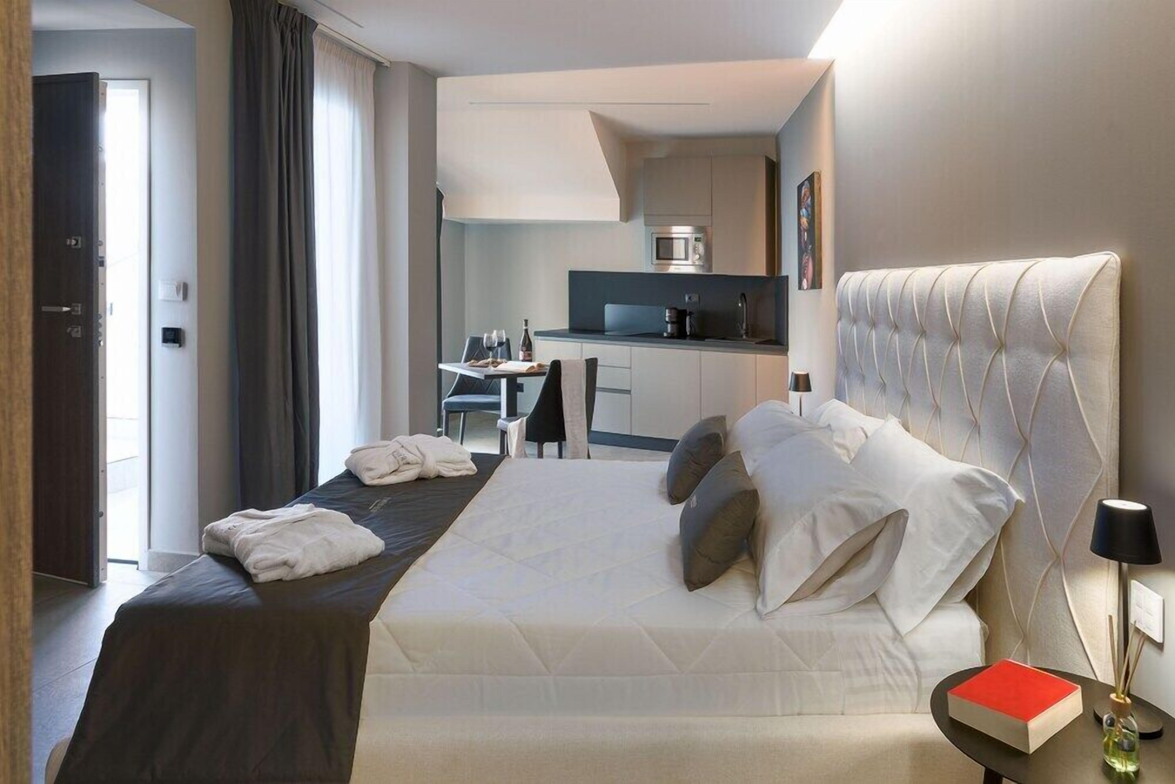 Quarto de hotel com cama, mesa de cabeceira com um livro, luminária e vista para a cozinha.