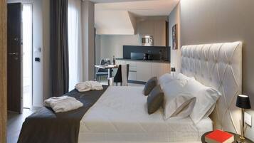 Quarto de hotel com cama, mesa de cabeceira com um livro, luminária e vista para a cozinha.