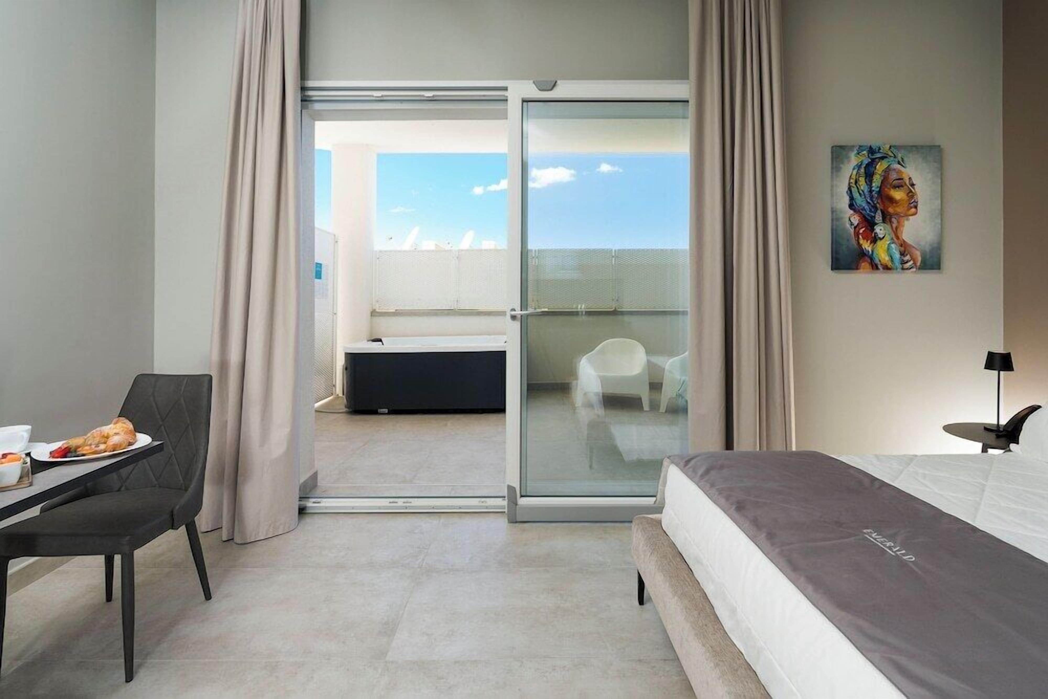 Quarto de hotel com cama, mesa de jantar com prato de comida, cadeira, quadro na parede e porta de vidro deslizante para uma varanda.