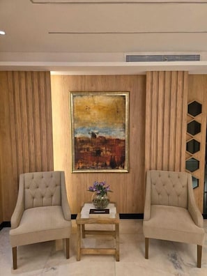 A room with two upholstered chairs, a small wooden table with a vase, and a large painting on the wall.