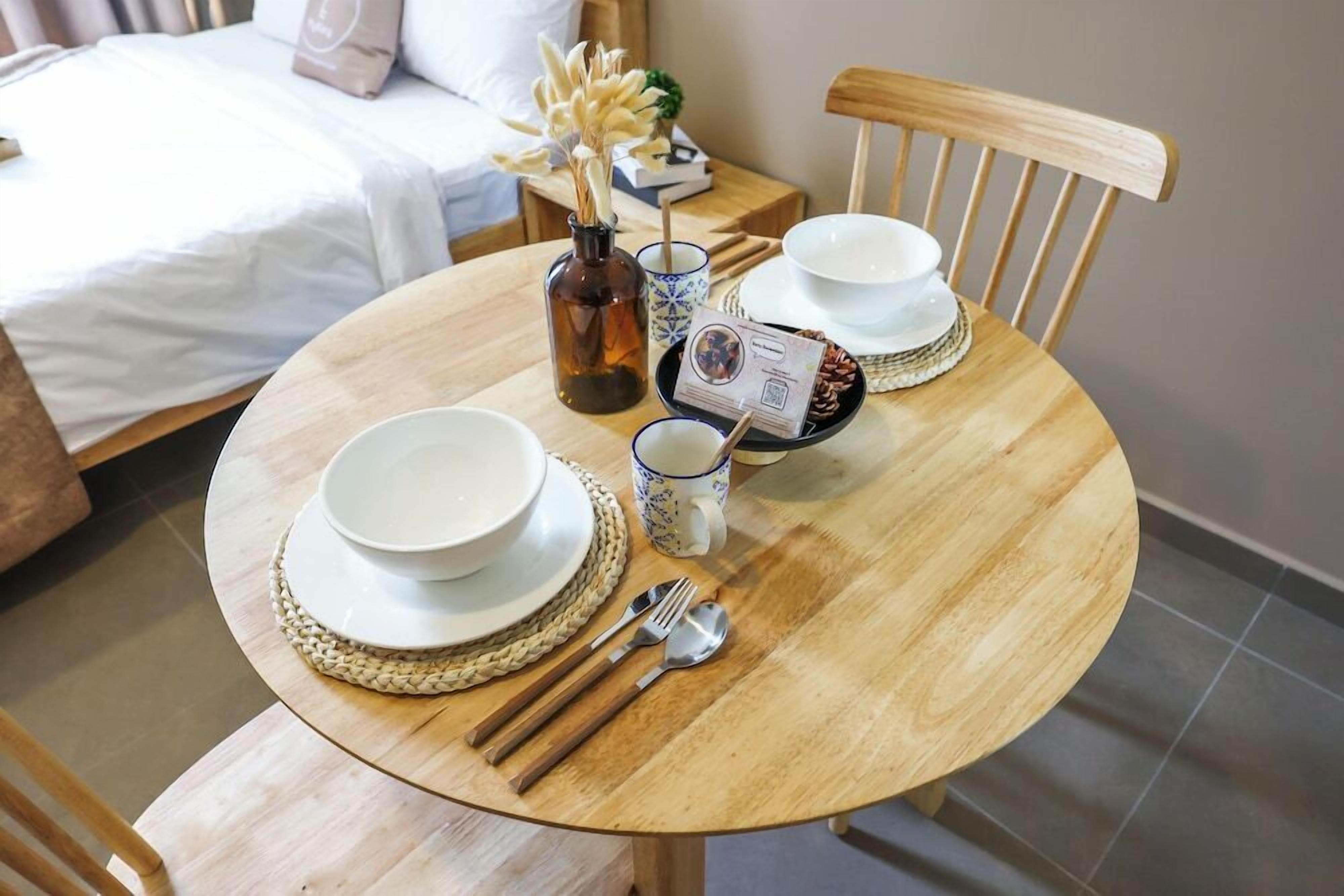A neatly arranged dining table with plates, bowls, chopsticks, and a mug, set in a room with a bed and a nightstand in the background.