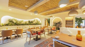 Ein Restaurant mit Holzbalkendecke, runden Tischen und Stühlen. Entlang der Wand stehen Pflanzen, und in der Ecke ein großer Kübel mit einer Pflanze.