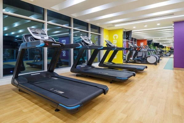 Un gimnasio con varias cintas de correr dispuestas en filas, ventanas amplias y suelo de madera.